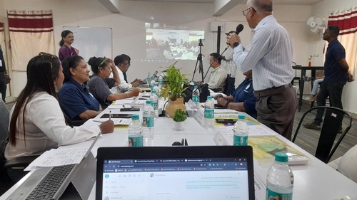 APM Meeting Session, the images depicts a photo taken during a meeting, there's a laptop screen and someone is talking on a microphone, there's a screen being projected on a wall