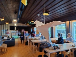 The photograph shows a dove made of paper, the dove is holding some sort of plant made with green paper, in the blurry background we can see a office with people sitting