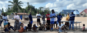 this photographs shows us a group of people, some of them are stading, some are sitting down on the ground, they seem to be waiting for something, some people are dressed ready to do a sports