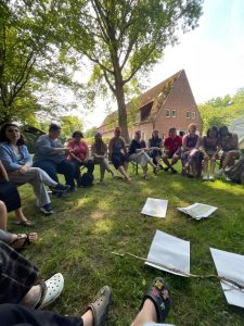 Photo taken in first person view, we can see a circle of people sitting in chairs, there's some papers in the grass, in the background we can see a house